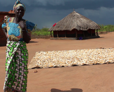 vlcsnap-2021-09-06-09h24m56s191 A woman standing with her arms crossed and one hand on her shoulder. Behind her is cassava being dried on the floor and a grassthatched house in the distance