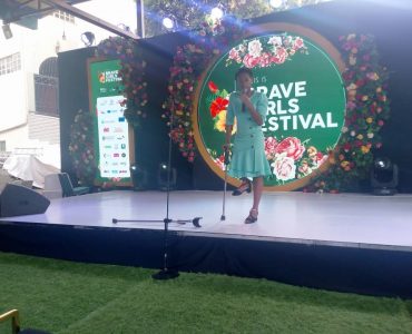 Maureen2 A young woman with a physical disability stands on a podium with a floral backdrop speaking into the microphone