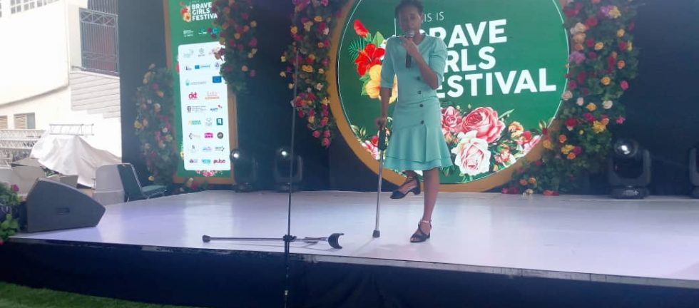 A young woman with a physical disability stands on a podium with a floral backdrop speaking into the microphone
