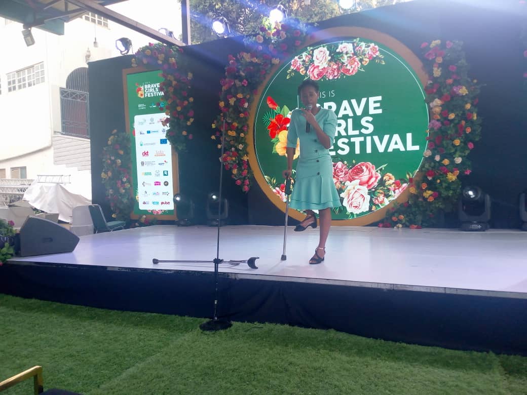 A young woman with a physical disability stands on a podium with a floral backdrop speaking into the microphone
